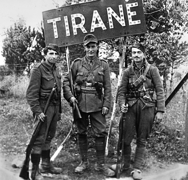 On November 28, 1944, the day Nazi rule ended in Albania, these guerrilla fighters pose under a sign that identifies their hometown. British efforts, led by the Special Operations Executive (SOE), to influence guerrilla operations in Albania met with only partial success at best.