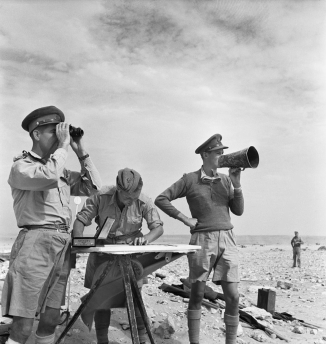 Manning the command post of the Royal Horse Artillery, officers use a map, megaphone, and field glasses to assess and redirect the accuracy of their fire against advancing Italian troops and tanks. The troop commander, left, is spotting the fall of the shells, while the gun position officer, right, gives the order to fire the heavy guns.