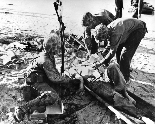 While a rifle is used to hang a bottle of plasma, a wounded Marine receives medical care on the sands of Tarawa.