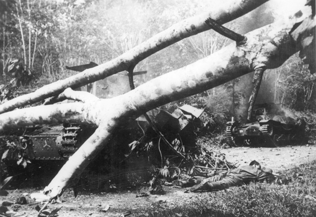 A dead Japanese crewman lies sprawled beside his tank while another wrecked tank smolders in the background. The armored vehicles were lost during a sharp clash with Australian troops who put up stiff resistance.
