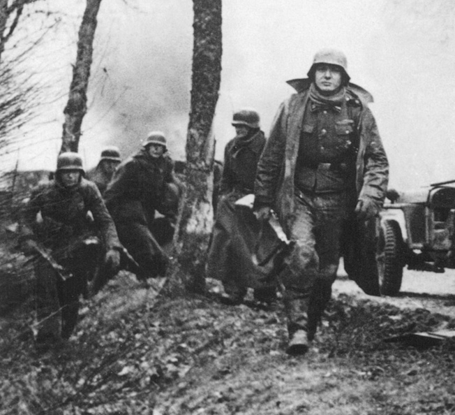 German troops stride past flaming American vehicles during the Battle of the Bulge. The soldier in the foreground carries a Mauser Kar 98 rifle, while the one at left holds a StG44 assault Rifle.