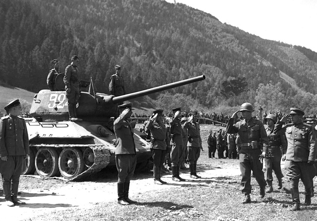 A detachment of Red Army Guards snaps to attention as a group of visiting Americans passes. A T-34 tank squats with the Soviets on a parade ground after the end of hostilities.