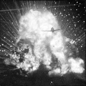 As a German truck loaded with ammunition explodes in a fireball, an American P-47 fighter flies through the flames, smoke, and debris.