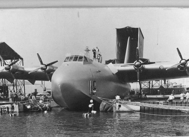 Hughes, on top of the fuselage, supervises operations to launch his plane from Long Beach. Note the specially constructed, climate-controlled hangar in the background, large enough to accommodate the massive tail.