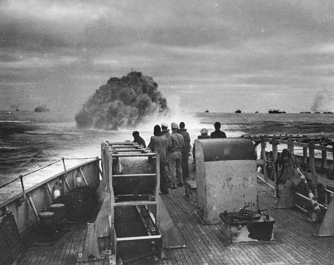 Crewmen aboard the U.S. Coast Guard Cutter Spencer watch the waters of the Atlantic Ocean brew up with the detonation of a depth charge. This photograph was taken while the Spencer was defending a trans-Atlantic convoy, visible in the background, against a German U-boat attack.